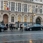 Façade grise de l'Hôtel de Ville sous un ciel couvert, voitures officielles et enquêteurs discrets portant des cartons de preuves.